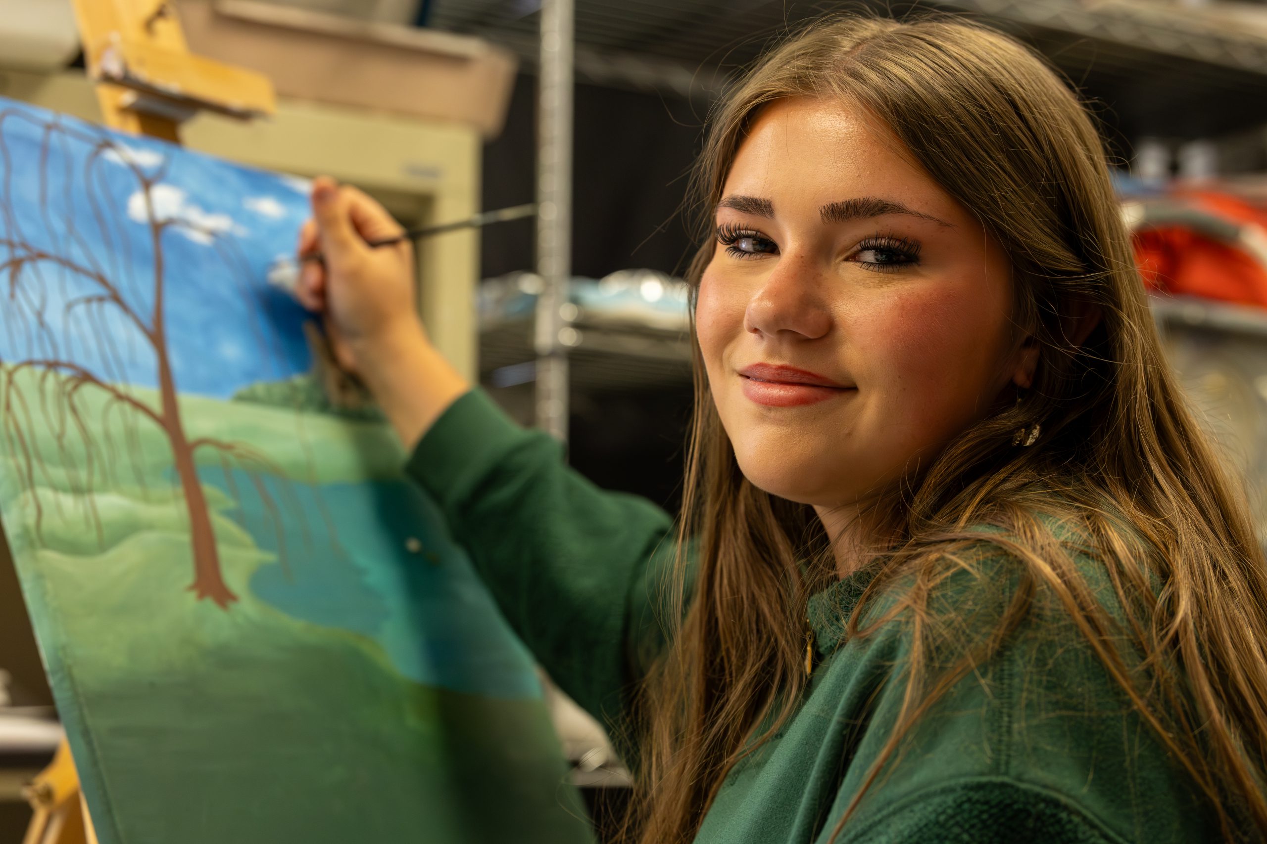 A high school student smiles at the camera while painting a tree