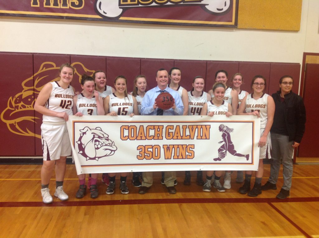 Coach Tom Galvin holds up a celebratory ball with his team posing behind a banner after his 350th win.