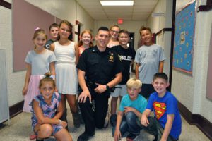 Officer in uniform on one knee surrounded by a dozen fourth grade students