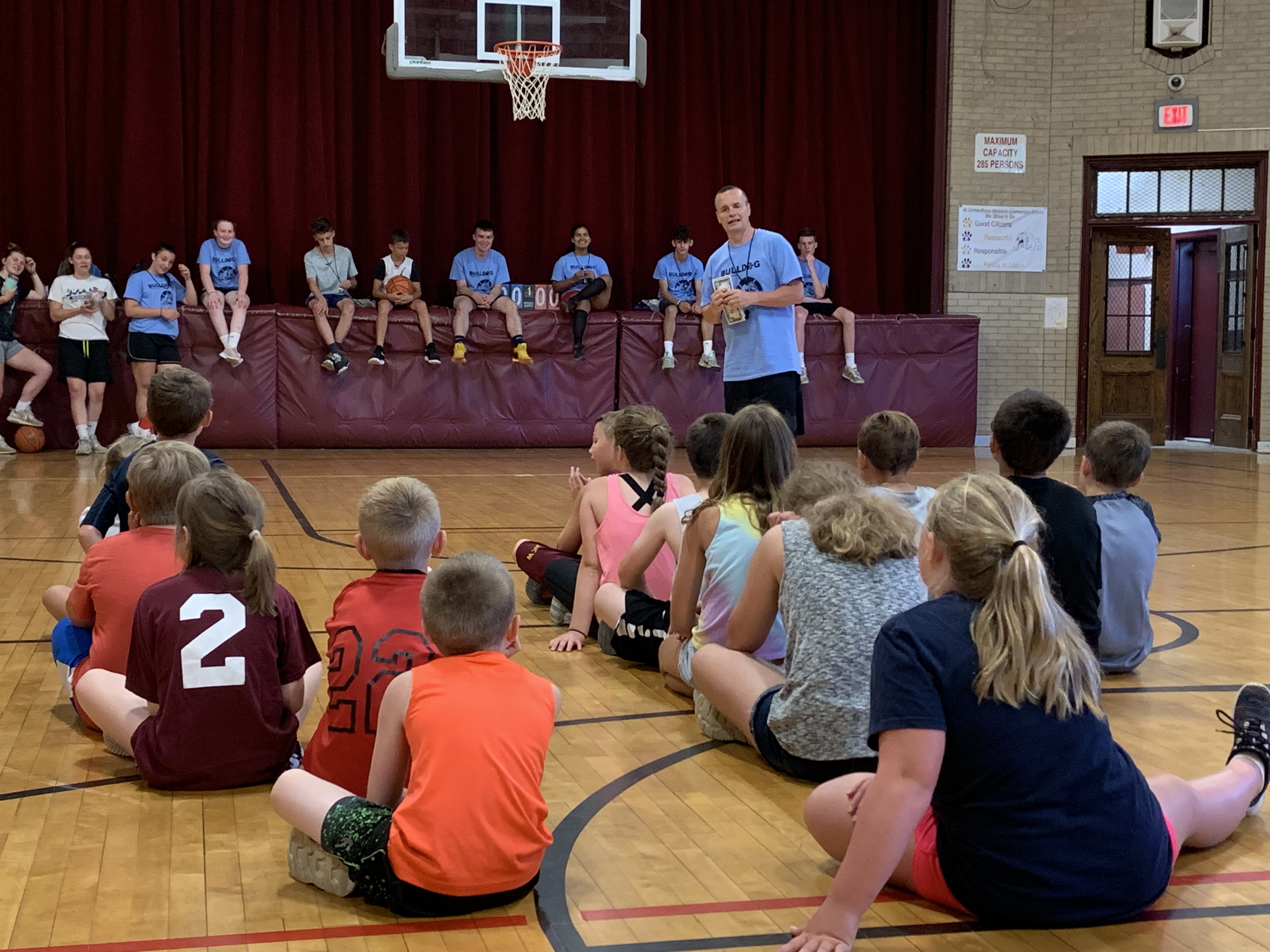 coach talks to group of students as they sit on a gym floor