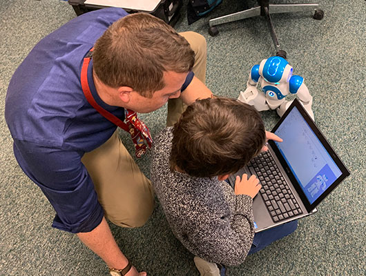 teacher and student work on laptop next to Natasha the robot