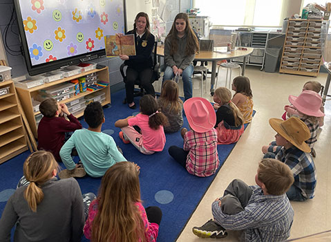 2 students seated reading a book to younger students sitting on chairs on floor