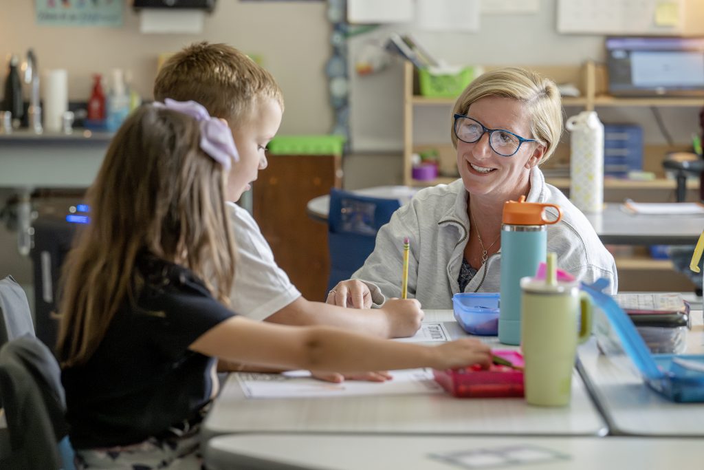 teacher smiling at young students and assisting them with writing