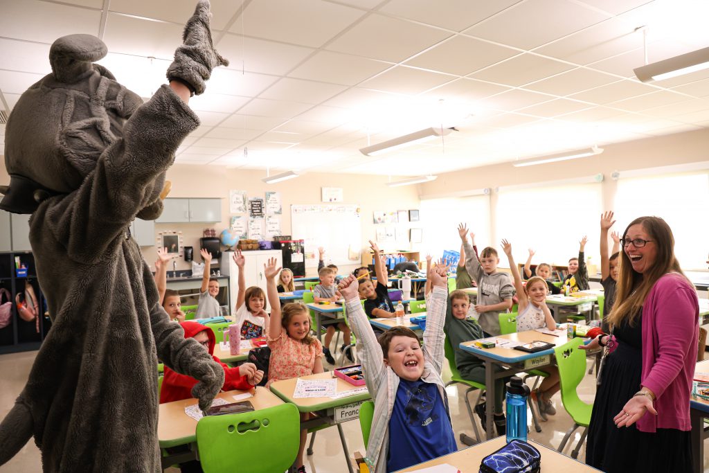 bernie the bulldog mascot and students in a classroom smiling and raising their hands with teacher standing in front of classroom laughing