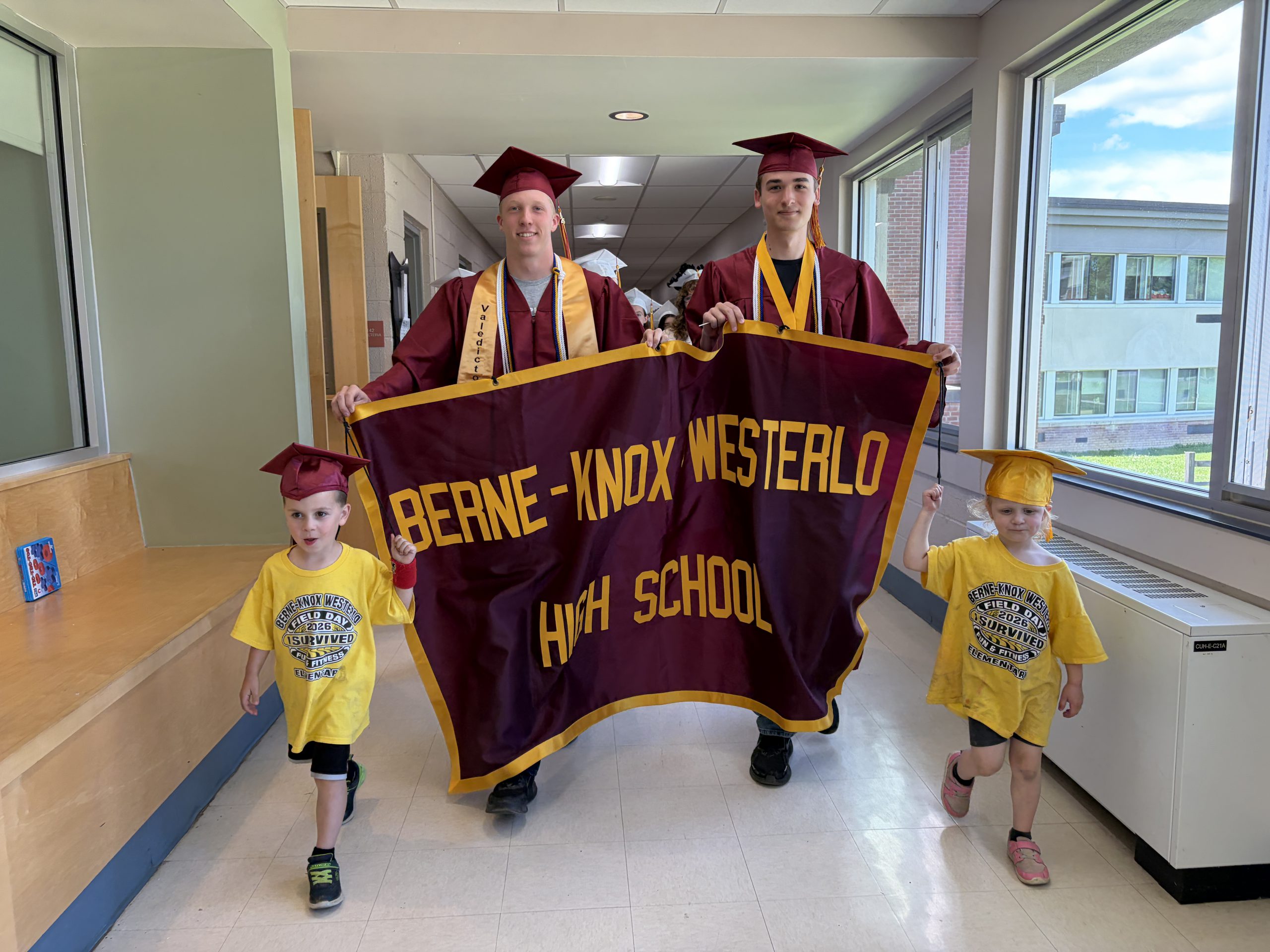 PreK students lead the senior graduation walkthrough while seniors hold a banner